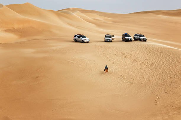 In the Siwa Oasis desert in Egypt, four off-road vehicles stand on golden dunes. A lone person walks toward them, leaving footprints across the vast, silent sand landscape.