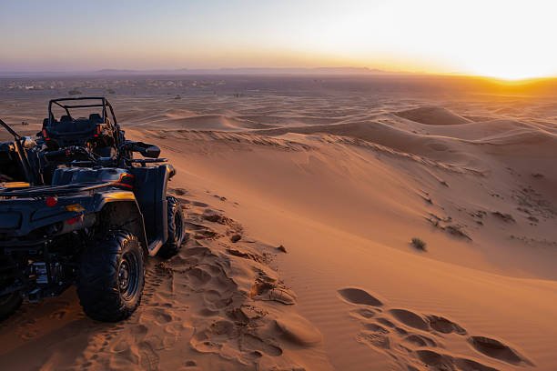 A dark green ATV, quad bike, and a UTV are parked on the edge of a large sand dune overlooking a vast desert landscape at sunset, with a remote village visible in the distance, Erg Chebbi, Sahara desert, in Hassilabied, junto a Merzouga, Marruecos