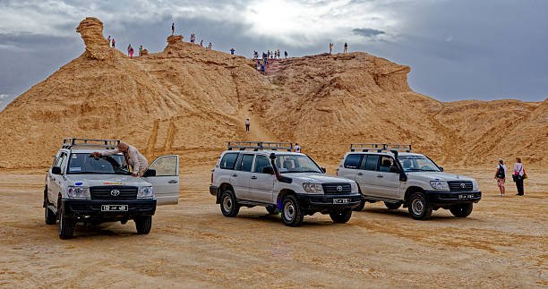 Three white 4x4 SUVs parked in the desert plains below the iconic Camel Rock formation with tourists climbing the peak in Ong Jmal, Tunisia. Captured on 08.10.2024